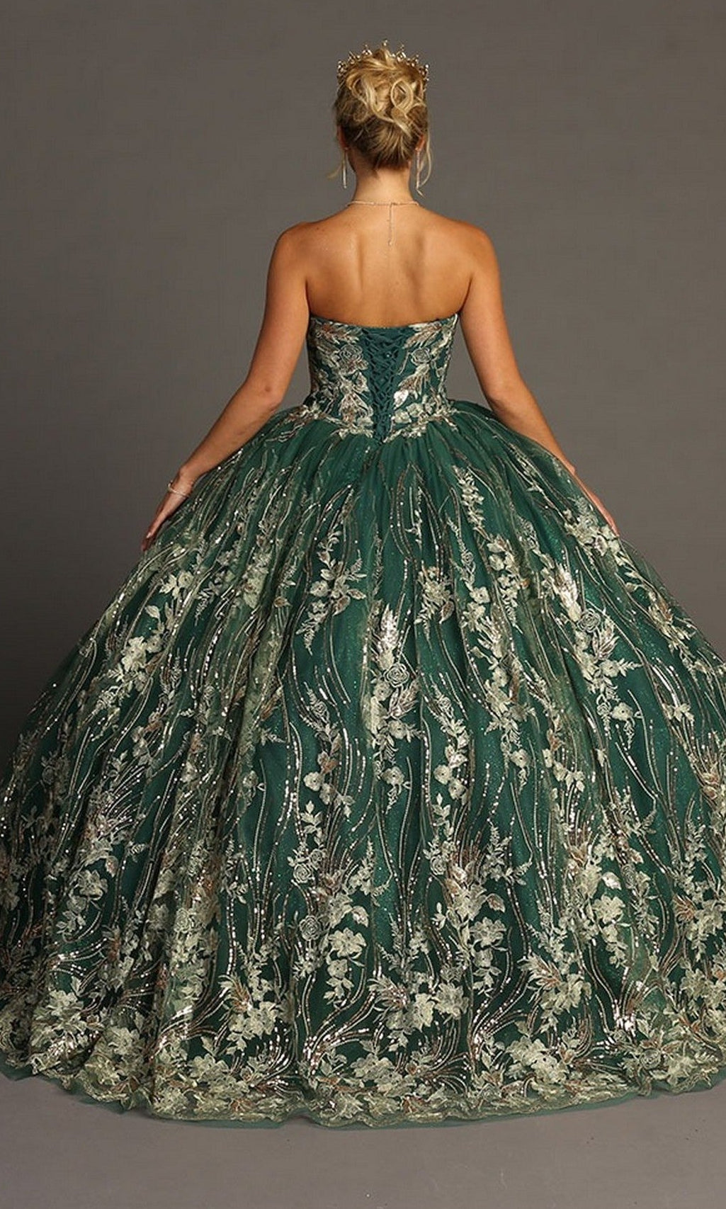 A woman wearing the Quinceanera Dress CHF3079 By Chicasโa strapless green ballgown with metallic floral embroideryโstands facing away from the camera against a plain gray background.